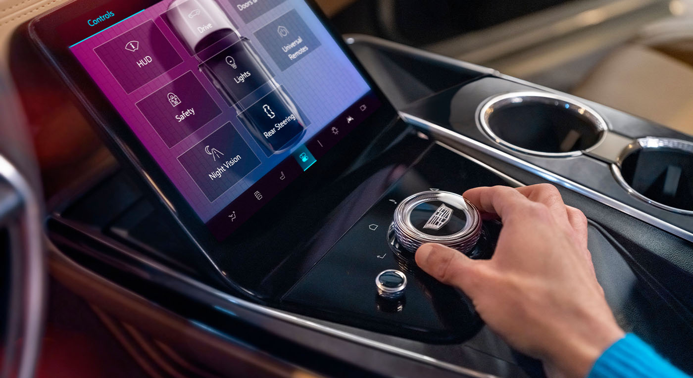 A hand adjusts a sleek, circular controller on Cadillac Escalade IQL a car's modern touchscreen dashboard, displaying safety, night vision, and lighting options, conveying innovation.